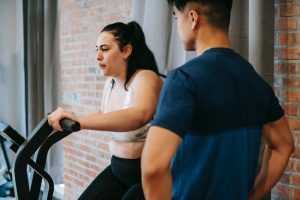 serious young asian male trainer looking at overweight woman exercising on gym bike