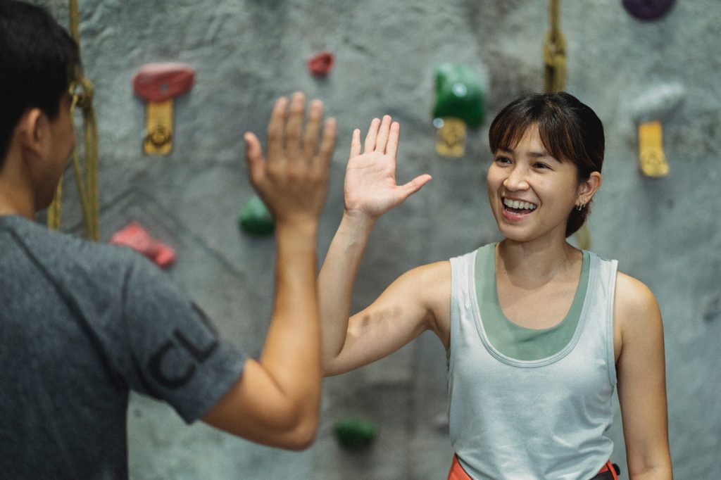 happy ethnic female climber with anonymous male friend giving high five in gym