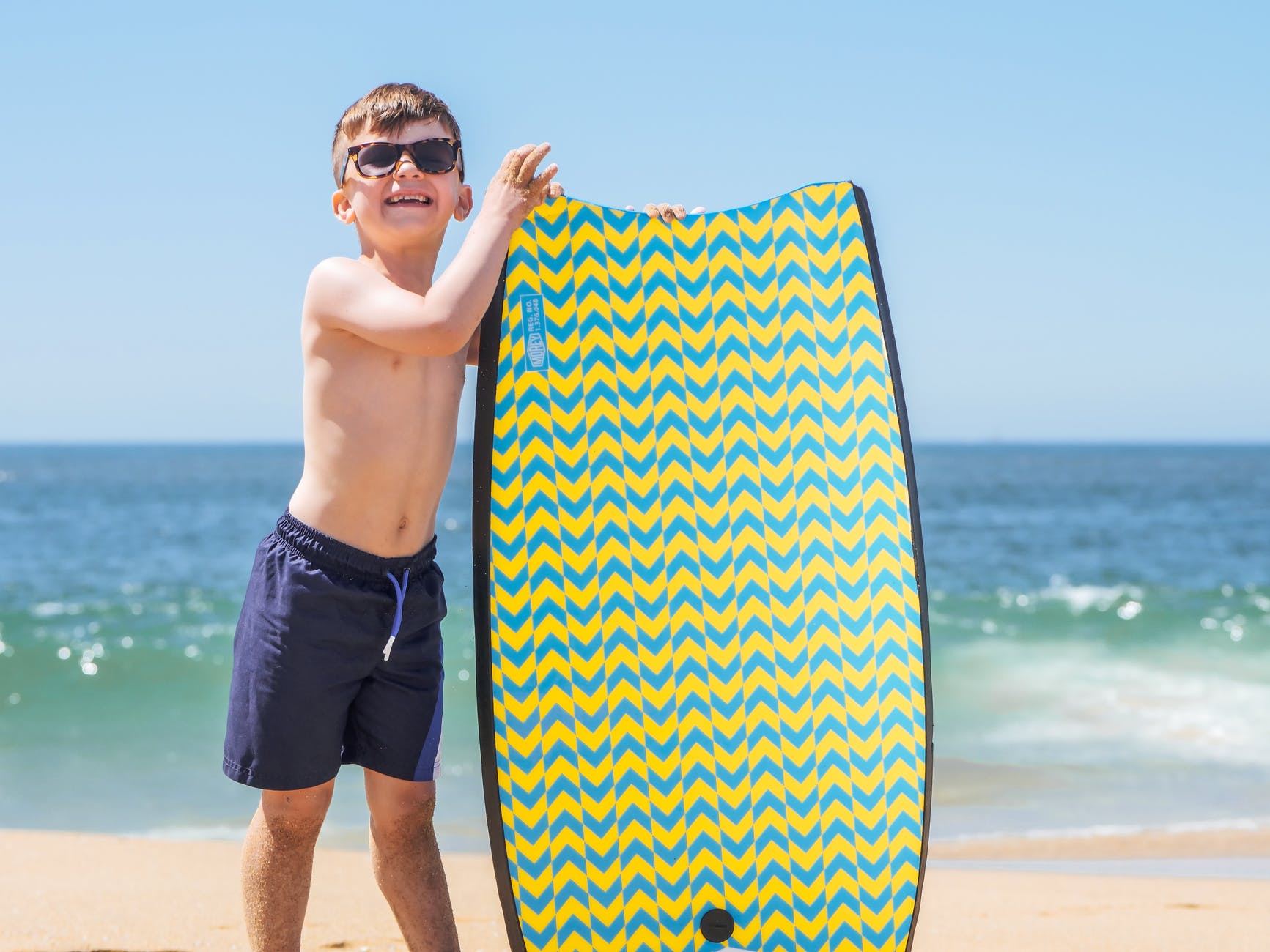 a boy wearing blue shorts and sunglasses holding a skimboard on the beach